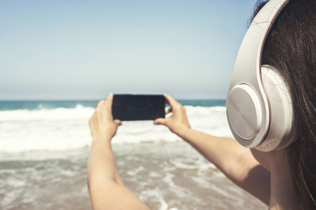 La mujer sola en la playa en los auriculares escuchan la música, tomando la foto del mar usando teléfono. Relajación femenina en vacaciones de verano. Vista trasera - Foto, Imagen