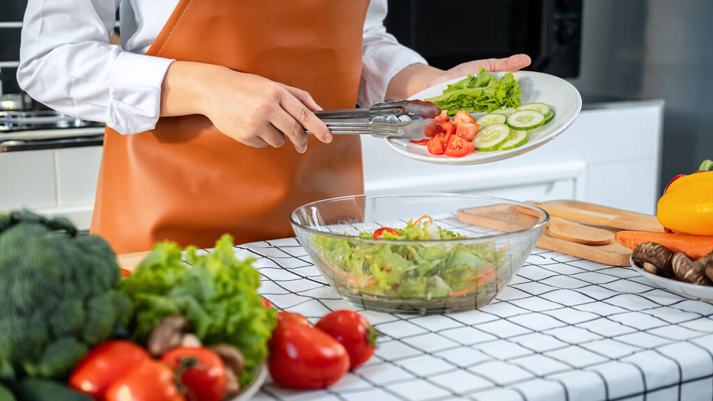 Ασιάτισσα νοικοκυρά κρατώντας πιάτο με μαρούλι, αγγούρι και ντομάτα και χρησιμοποιώντας λαβίδες λαχανικών σε μπολ για να αναμιγνύει σαλάτα για μεσημεριανό ενώ φοράει ποδιά και στέκεται στο μαγείρεμα υγιεινό γεύμα στην κουζίνα - Φωτογραφία, εικόνα