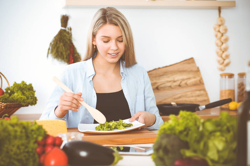 Приваблива молода блондинка смакує новий рецепт смачної салатної суміші, сидячи за столом на кухні
. - Фото, зображення