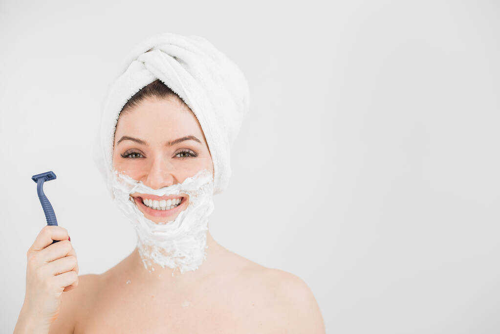 Femme caucasienne joyeuse avec une serviette sur la tête et mousse à raser sur son visage tient un rasoir sur un fond blanc - Photo, image