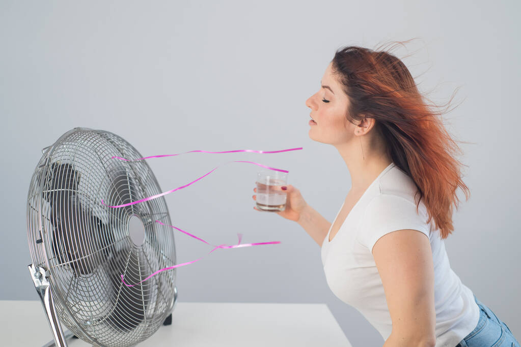 Uma mulher caucasiana ruiva se acalma junto ao ventilador elétrico e bebe uma bebida gelada. Controle do clima no apartamento - Foto, Imagem