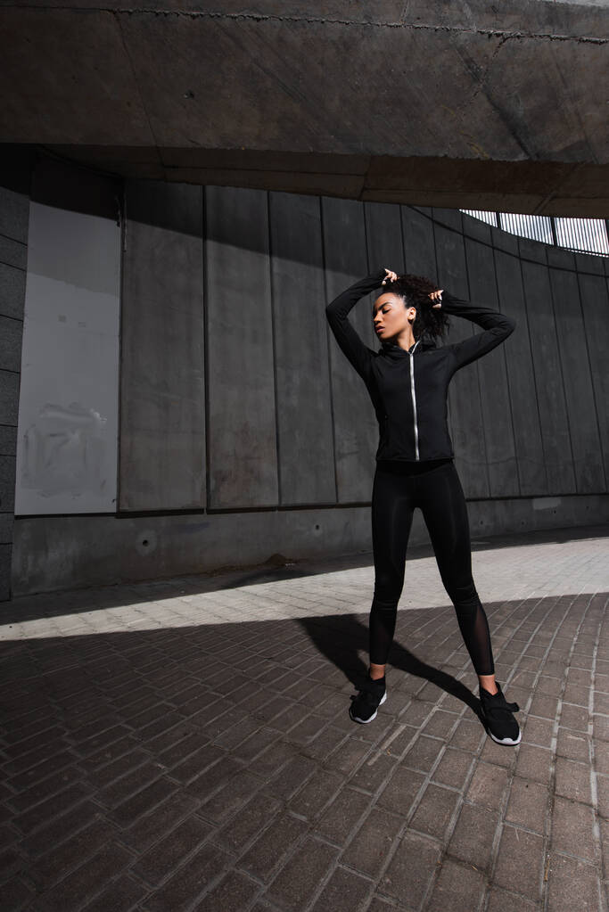 Zijaanzicht van Afro-Amerikaanse sportvrouw aanpassen haar op stedelijke straat  - Foto, afbeelding