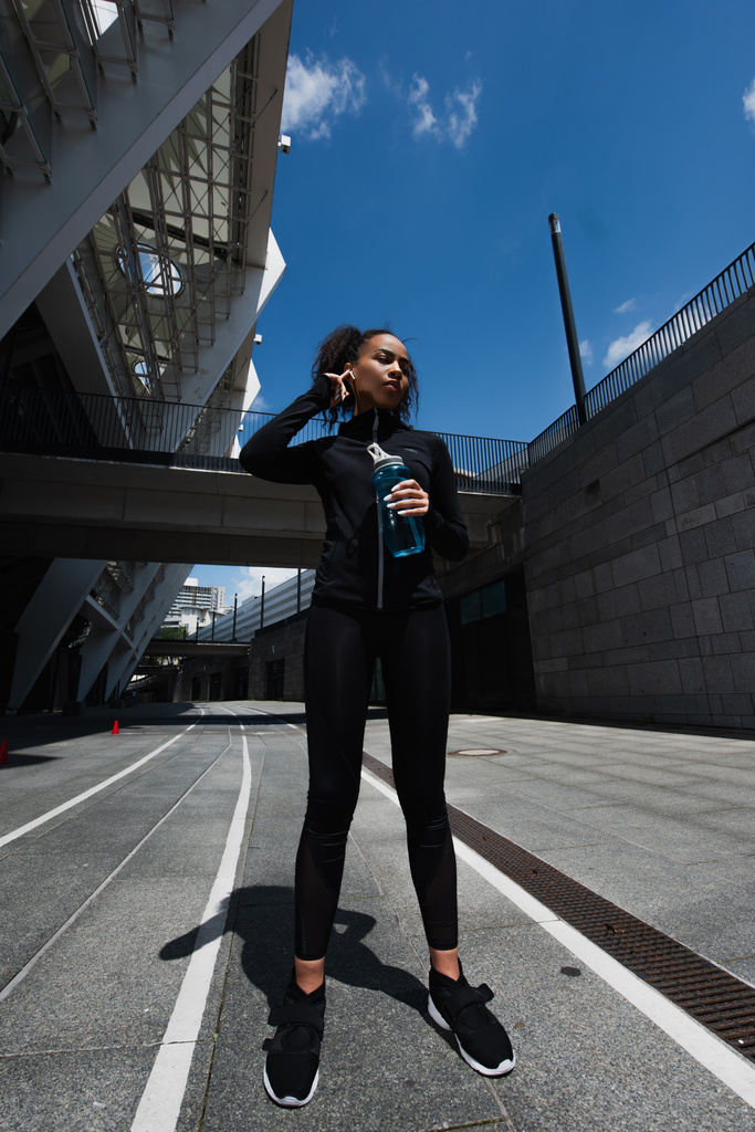 Афроамериканська спортсменка в навушниках, що тримає спортивну пляшку на міській вулиці.  - Фото, зображення