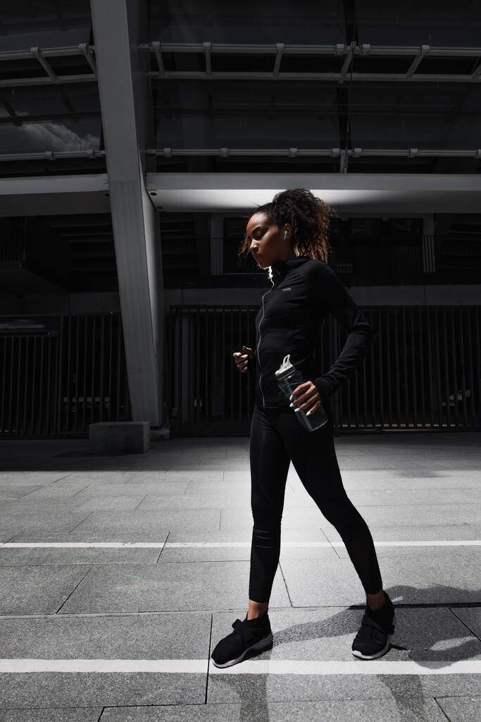 Boční pohled na africké americké sportovkyně ve sluchátku drží sportovní láhev na městské ulici ve večerních hodinách  - Fotografie, Obrázek