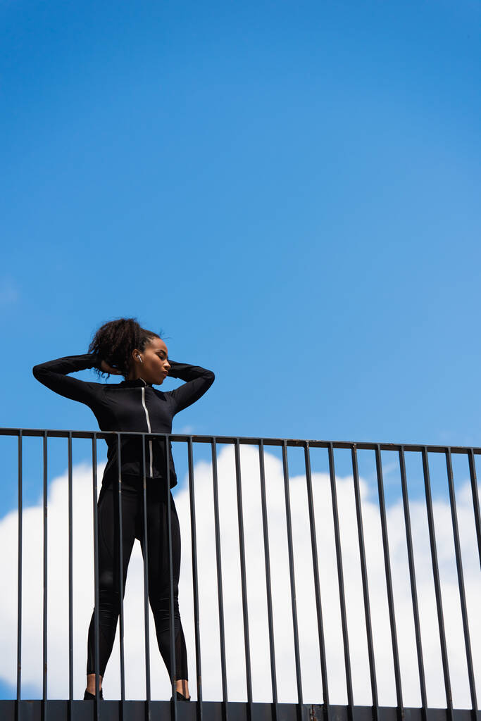 Άποψη χαμηλής γωνίας της Αφροαμερικανής αθλήτριας σε ακουστικό στέκεται με κλειστά μάτια στη γέφυρα  - Φωτογραφία, εικόνα
