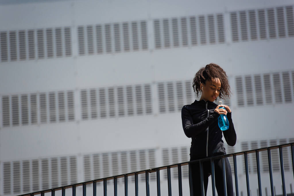 Африканская спортсменка в наушниках держит спортивную бутылку на мосту на городской улице  - Фото, изображение