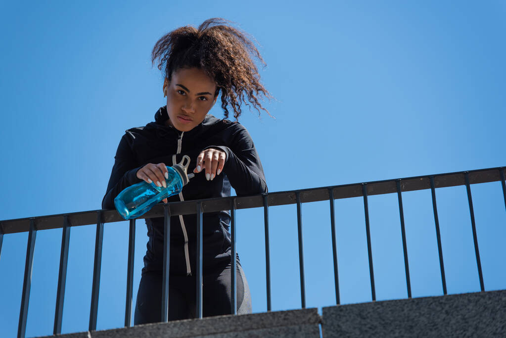 Tiefansicht einer jungen afrikanisch-amerikanischen Sportlerin mit Kopfhörer, die eine Sportflasche mit blauem Himmel im Hintergrund hält  - Foto, Bild