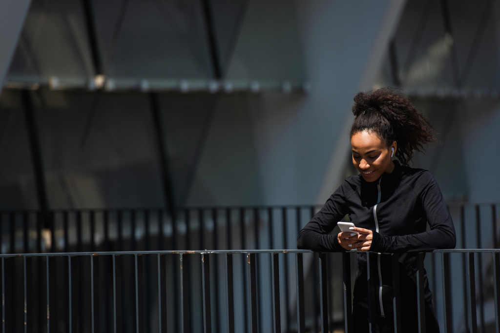 Mosolygó afro-amerikai sportos nő fülhallgató segítségével mobiltelefon a városi utcán  - Fotó, kép