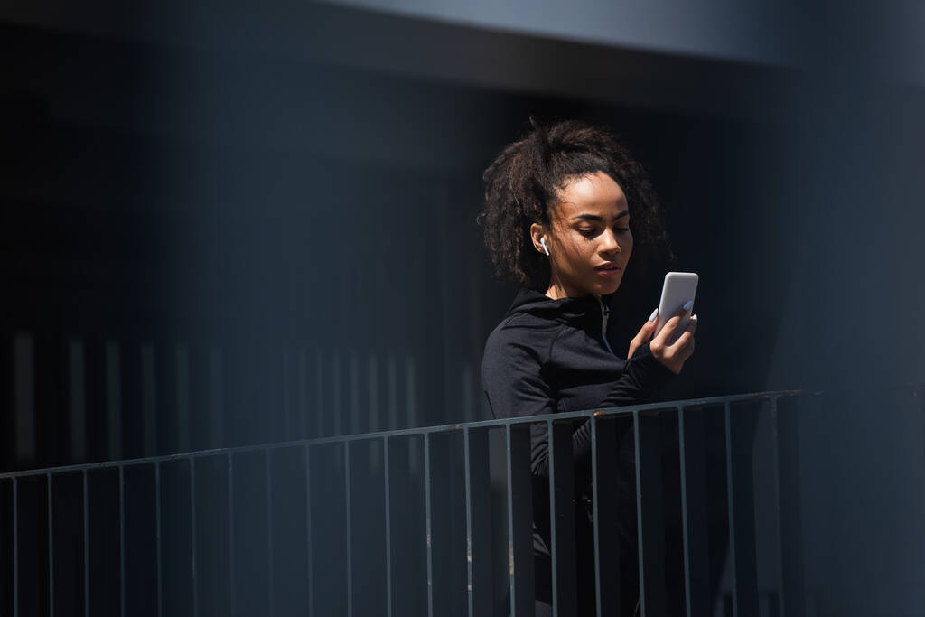 Junge afrikanisch-amerikanische Sportlerin im Kopfhörer mit Handy im Freien  - Foto, Bild