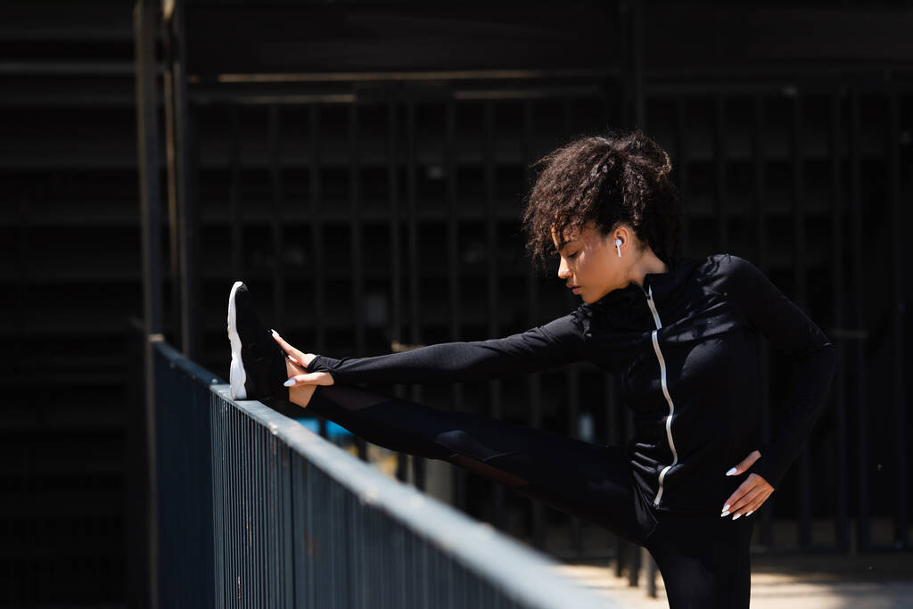 都市の通りに橋の近くにストレッチワイヤレスイヤホンでアフリカ系アメリカ人スポーツ女性の側面図  - 写真・画像