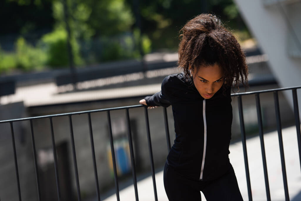 Joven deportista afroamericana escuchando música en auriculares y calentándose en la calle urbana  - Foto, Imagen