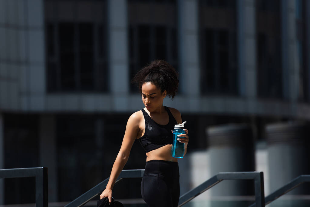 fit afrikaanse amerikaanse sportvrouw holding sport fles in de buurt reling buiten  - Foto, afbeelding