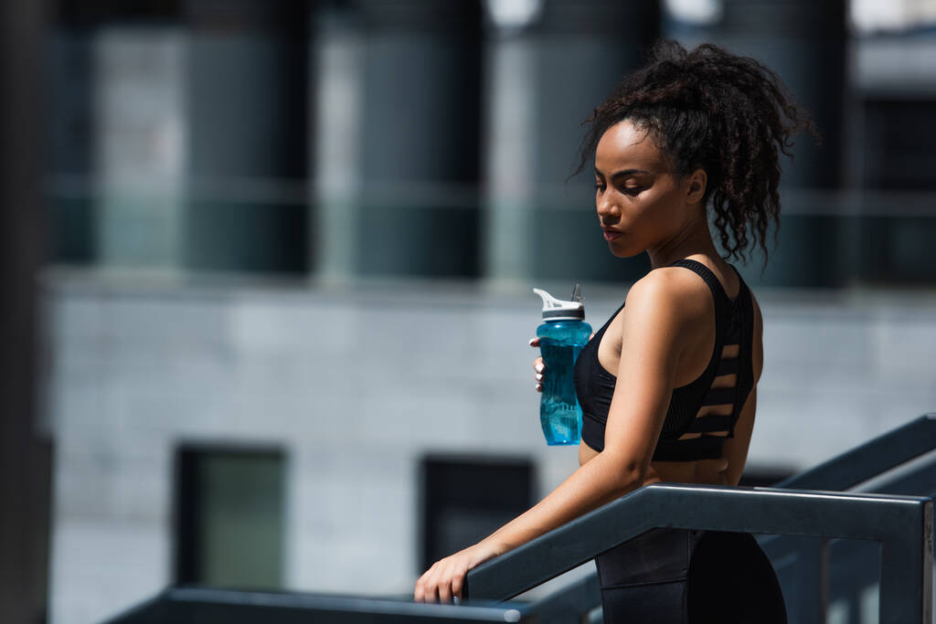 Afroamerikanische Sportlerin in schwarzem Top mit Sportflasche am Geländer im Freien  - Foto, Bild