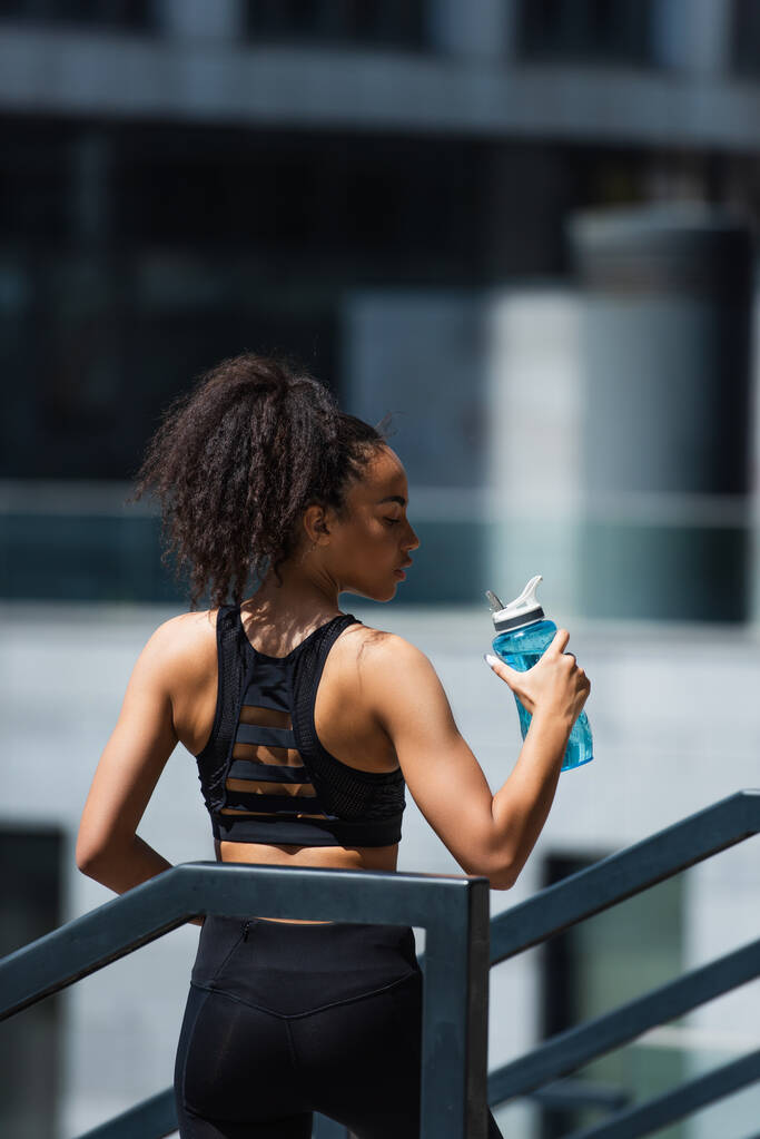 Vista lateral de bastante afroamericana deportista sosteniendo botella de deportes con agua en la calle urbana  - Foto, Imagen