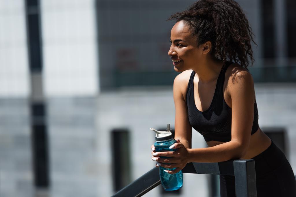 Jolie sportive afro-américaine avec bouteille de sport souriante près de la rampe à l'extérieur  - Photo, image