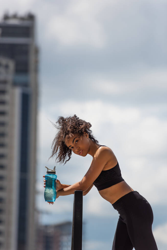 Fit afrikanisch-amerikanische Sportlerin hält Sportflasche in der Hand und blickt in die Kamera  - Foto, Bild