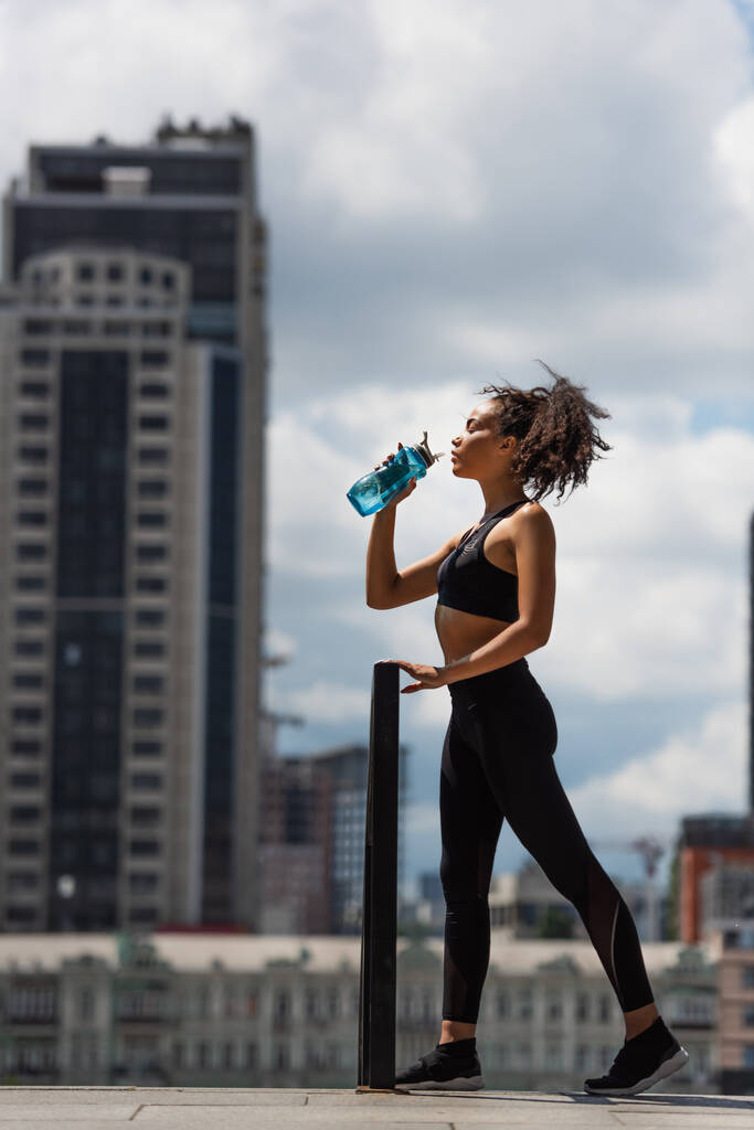 Seitenansicht einer afrikanisch-amerikanischen Sportlerin, die Wasser aus einer Sportflasche auf einer städtischen Straße trinkt  - Foto, Bild