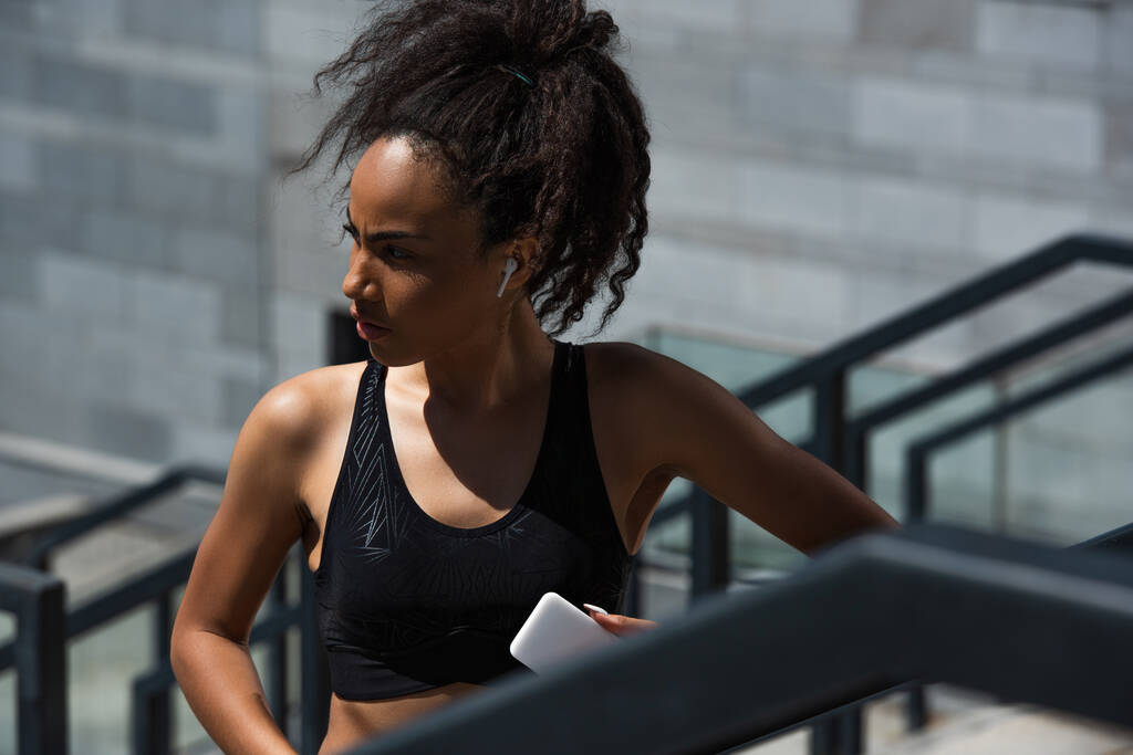 Africká americká sportovkyně ve sluchátku drží mobilní telefon venku  - Fotografie, Obrázek