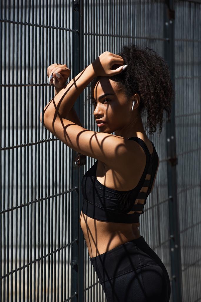 Donna afroamericana in auricolare e abbigliamento sportivo in piedi vicino alla recinzione  - Foto, immagini