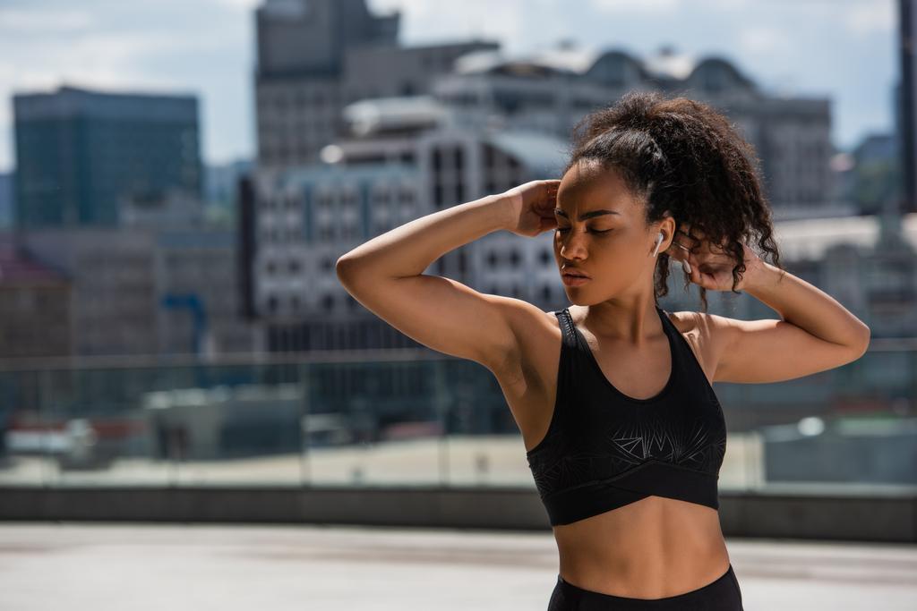 African american sportswoman in earphone stretching arms on urban street  - Photo, Image