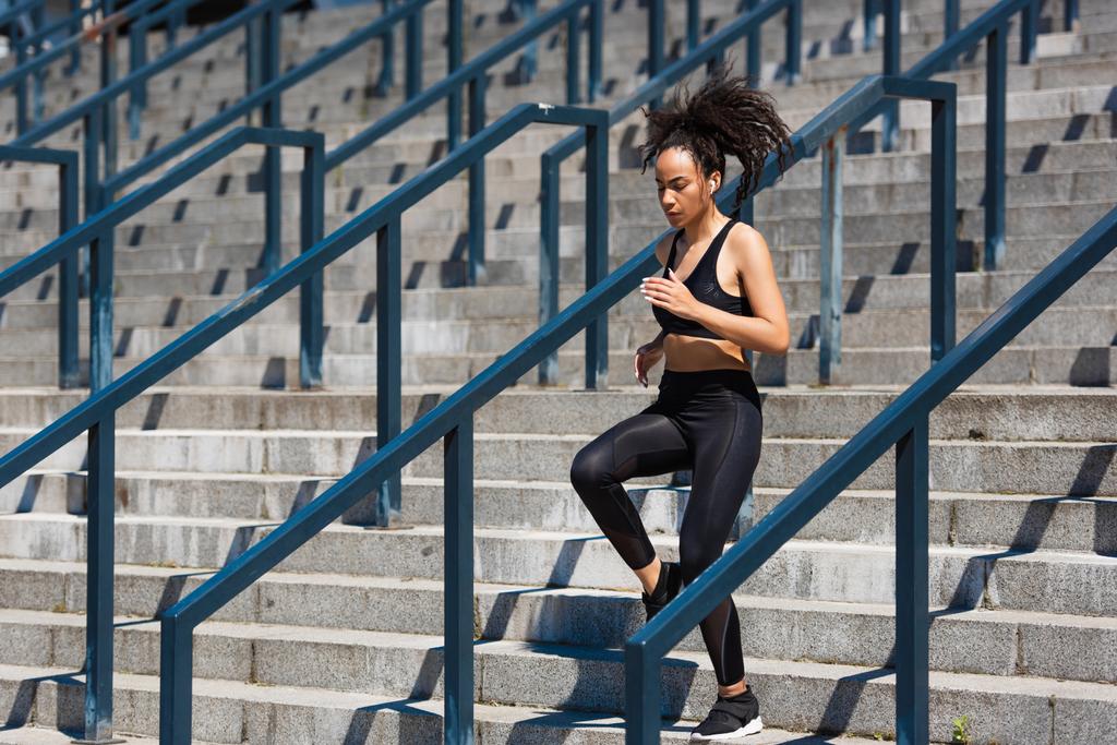 Afro-Amerikaanse sportvrouw in oortelefoon loopt op trappen in de buurt reling op stedelijke straat  - Foto, afbeelding