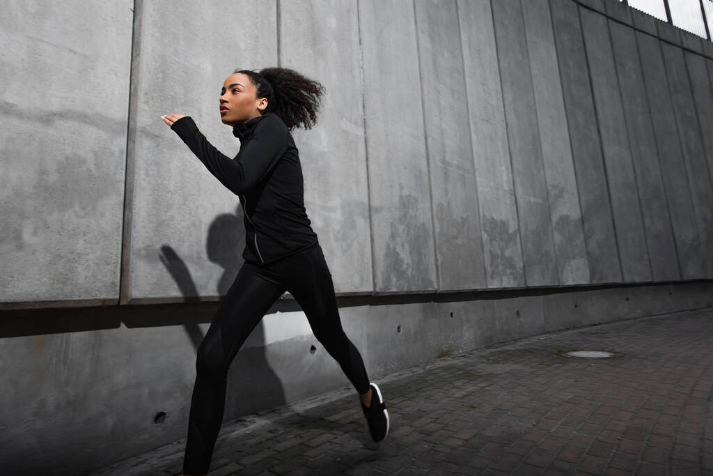 Νεαρή Αφροαμερικανίδα αθλήτρια που τρέχει κοντά σε τσιμεντένιο τοίχο με σκιά στον αστικό δρόμο  - Φωτογραφία, εικόνα