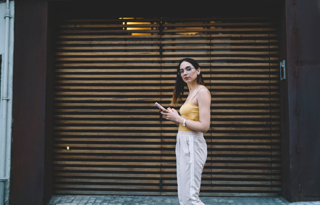 Poloviční portrét atraktivní cestovní blogger v brýlích procházky na městském prostředí a při pohledu na kameru, krásná hipster dívka s digitálním mobilem gadget návštěvě moderního města na dovolené - Fotografie, Obrázek