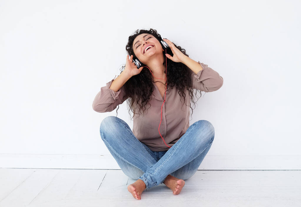 Νεαρή χαρούμενη Αφρο-Αμερικανίδα γυναίκα που φοράει ακουστικά κάθεται στο πάτωμα και ακούει μουσική, ενθουσιασμένη γυναίκα μικτής φυλής - Φωτογραφία, εικόνα
