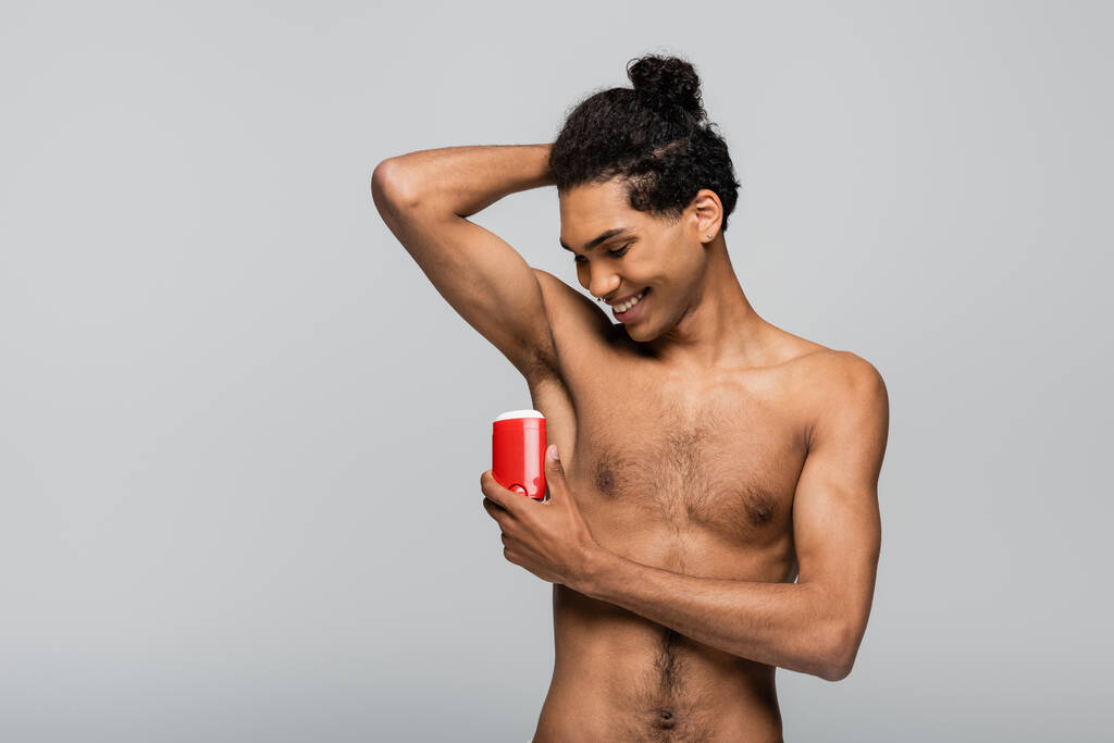 shirtless african american man smiling while applying dry antiperspirant isolated on grey - Photo, Image