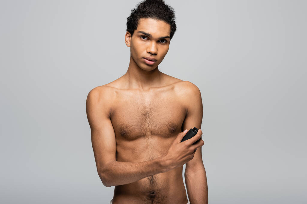shirtless, young african american man looking at camera while spraying antiperspirant isolated on grey - Photo, Image