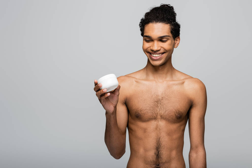 веселый, без рубашки африканский мужчина держит контейнер косметического крема изолирован на серый - Фото, изображение