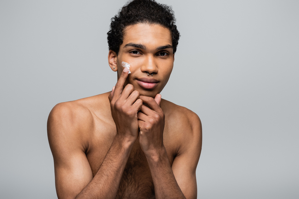 young african american man apply face cream while smiling at camera isolated on grey - Photo, Image
