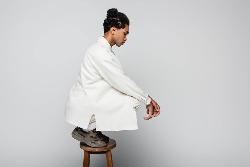 side view of african american man on high stool in white jacket and sneakers on grey - Photo, Image