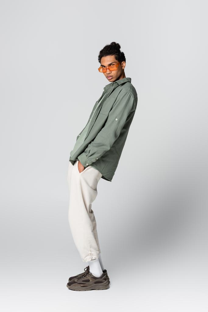 trendy african american guy looking at camera while holding hand in pocket on grey background - Photo, Image