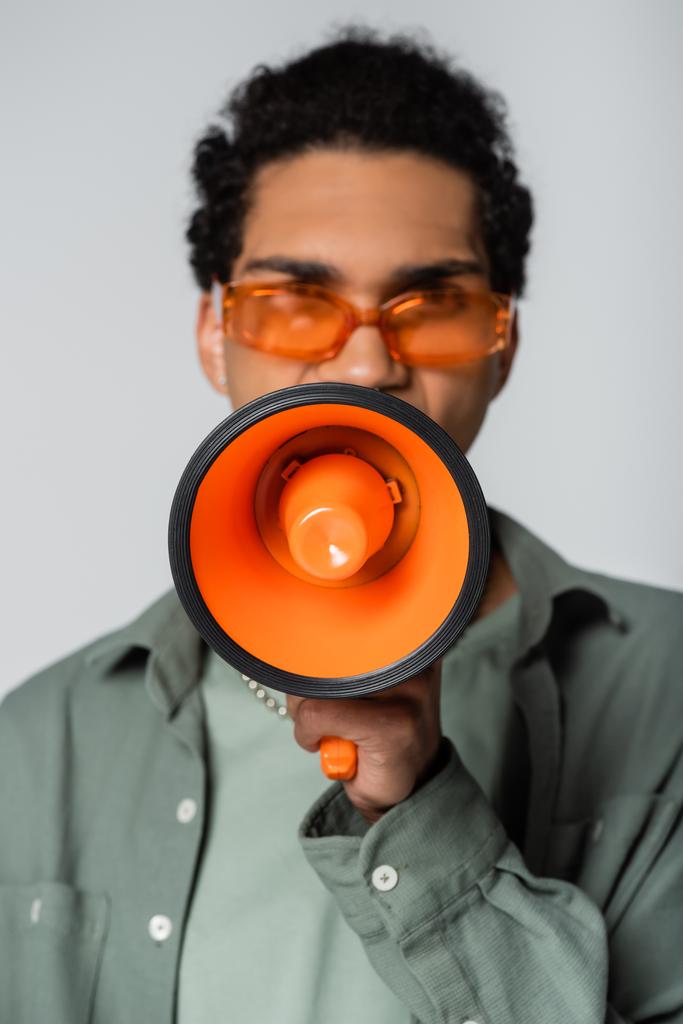 vista frontal del hombre afroamericano borroso en gafas gritando en altavoz naranja aislado en gris - Foto, imagen