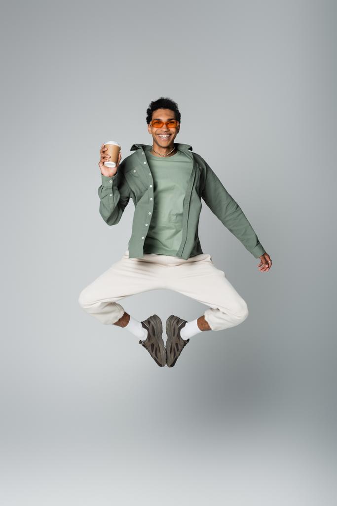 hombre afroamericano excitado en ropa de moda saltando con taza de papel aislado en gris - Foto, Imagen