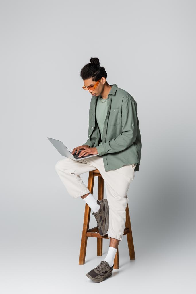 homme afro-américain à la mode tapant sur un ordinateur portable assis sur un tabouret haut sur fond gris - Photo, image
