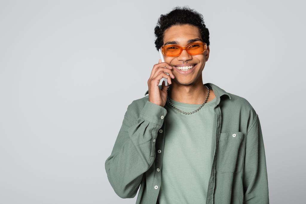 alegre afroamericano hombre sonriendo a la cámara mientras habla en el teléfono celular aislado en gris - Foto, imagen