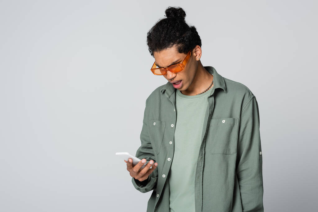 sorprendido hombre afroamericano en gafas naranjas mirando el teléfono celular aislado en gris - Foto, imagen