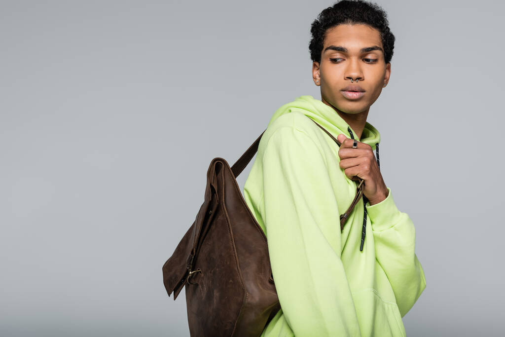 stylowy Afroamerykanin w zielonej bluzie, ze skórzanym plecakiem odizolowanym na szaro - Zdjęcie, obraz