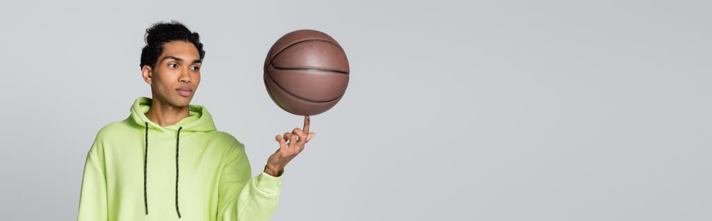 νεαρός Αφροαμερικάνος με πράσινη κουκούλα που παίζει μπάσκετ απομονωμένος σε γκρι λάβαρο. - Φωτογραφία, εικόνα