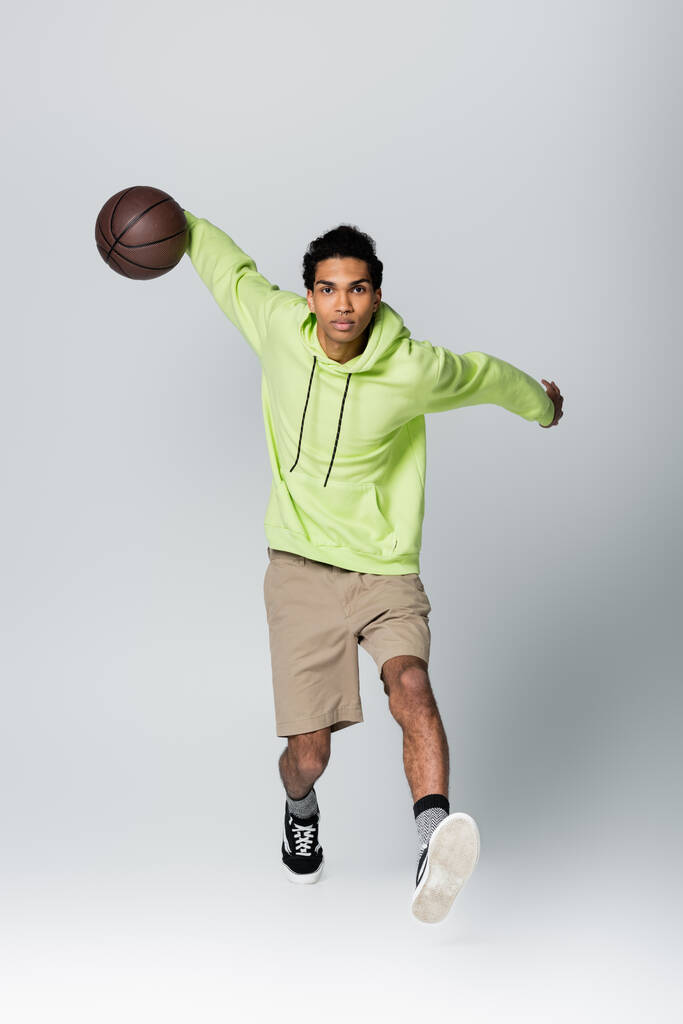 Gri arka planda basketbol oynayan Afro-Amerikan erkeğinin tam boy görüşü - Fotoğraf, Görsel