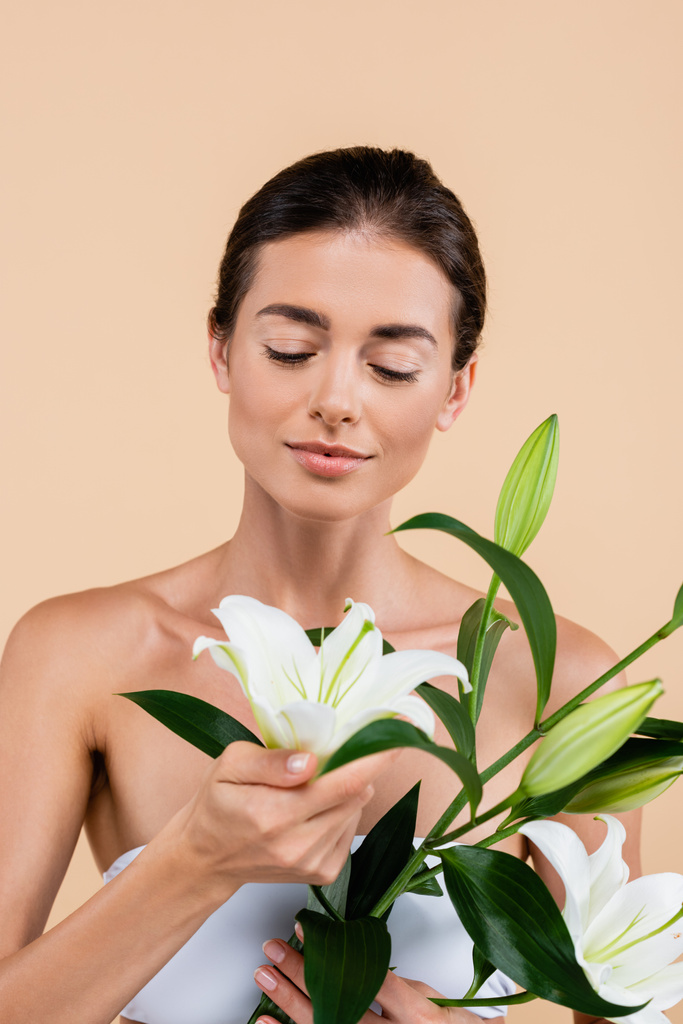 charming woman with naked shoulders looking at white lily flowers isolated on beige, beauty concept - Foto, Imagem