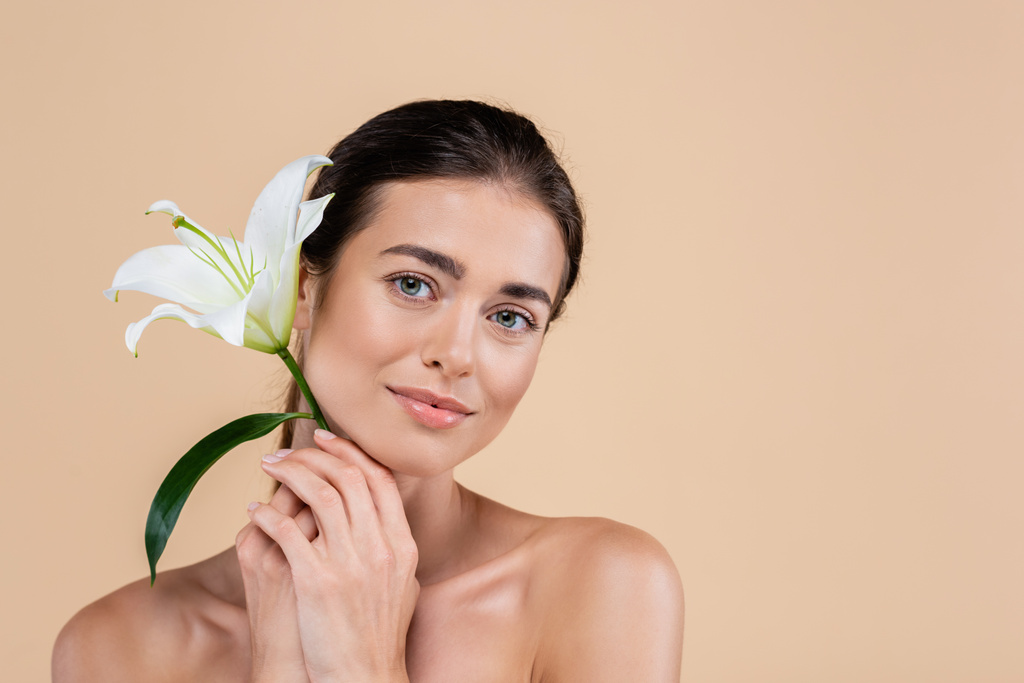 young woman with naked shoulders holding lily flower near face isolated on beige, beauty concept - Φωτογραφία, εικόνα