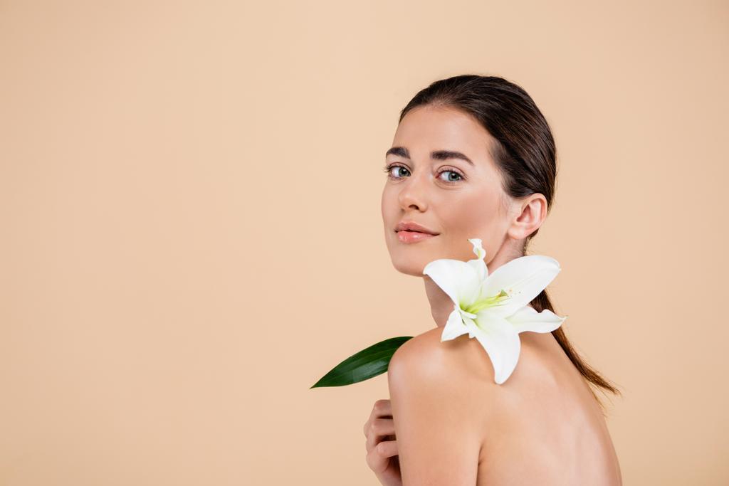 smyslná žena s bílou lilií květin na nahém rameni při pohledu na kameru izolované na béžové, krása koncept - Fotografie, Obrázek