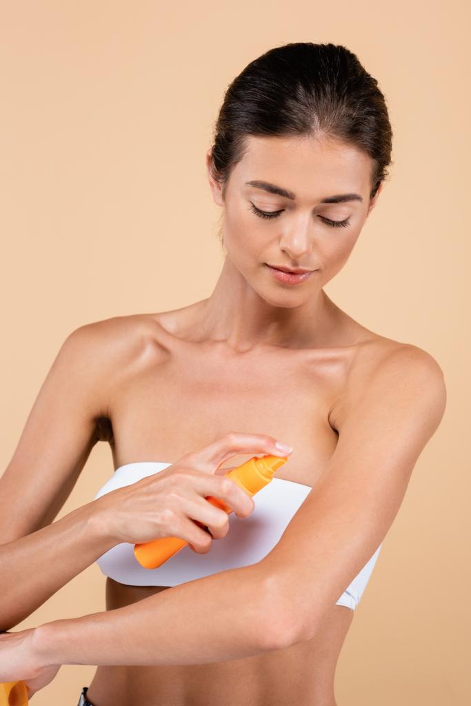 mooie vrouw met naakt schouders sproeien zonnebrandcrème op arm geïsoleerd op beige - Foto, afbeelding