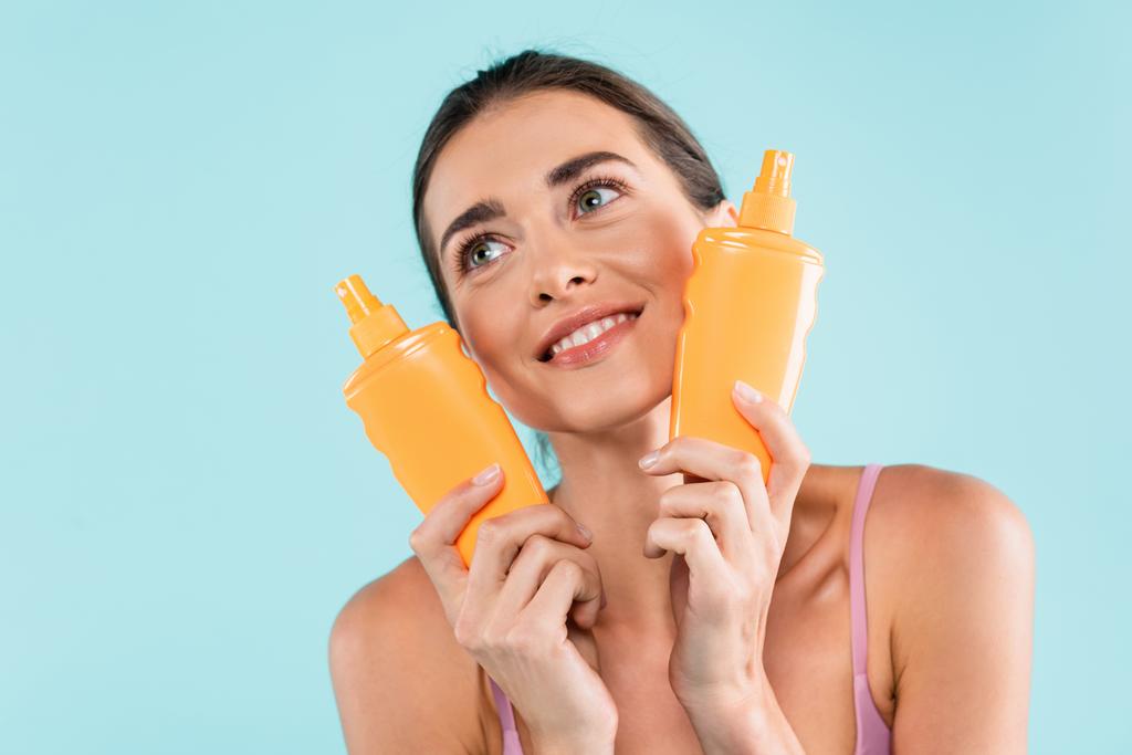 happy woman with bottles of sunscreen looking away isolated on blue - Foto, Imagem