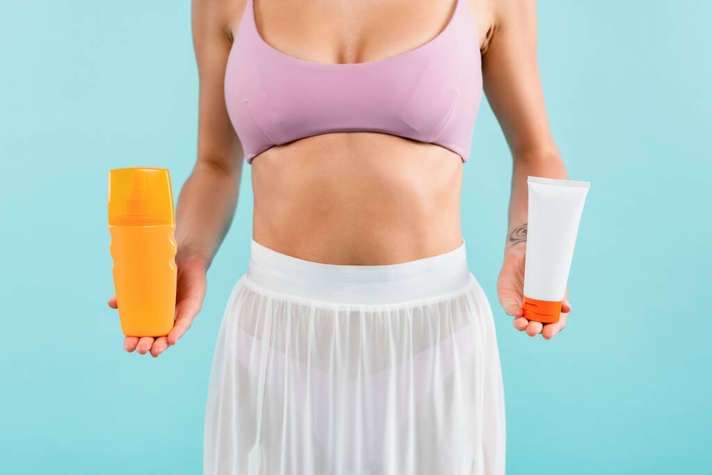 cropped view of woman in swimsuit and pareo holding tube and bottle of sunscreen isolated on blue - Photo, Image