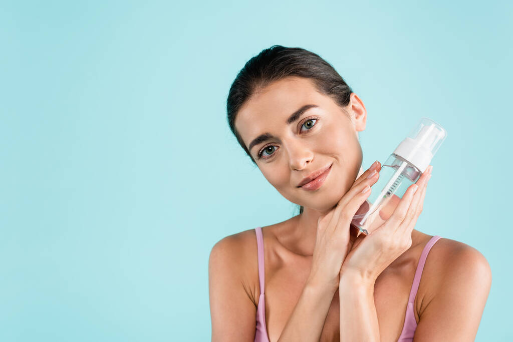 pretty woman holding bottle of face tonic while smiling isolated on blue - Foto, Imagem