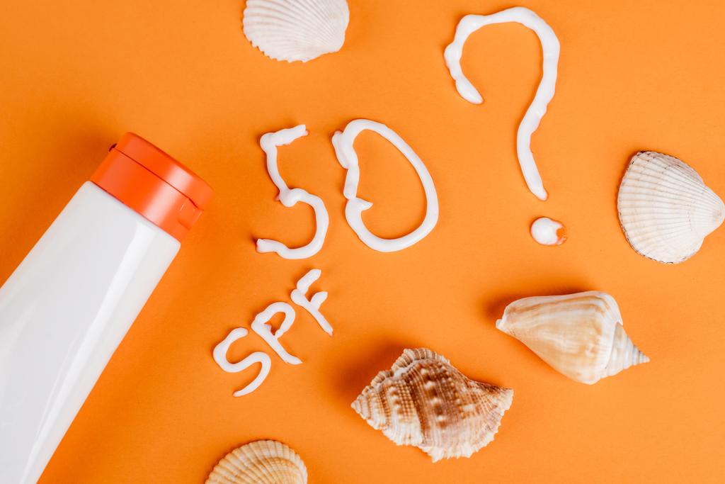 top view of sunscreen near seashells, spf lettering and number fifty with question mark on orange surface - Φωτογραφία, εικόνα